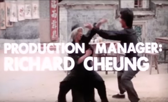 Two martial artists are engaged in combat in an outdoor setting with traditional buildings in the background. The text 'PRODUCTION MANAGER: RICHARD CHEUNG' is overlaid in large white letters across the image. Some of this text is hard to read due to the white text blending in with some of the light gray buildings in the background.